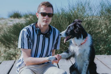 Kulaklıklı bir adam köpeğiyle sahildeki köprüde oturuyor.