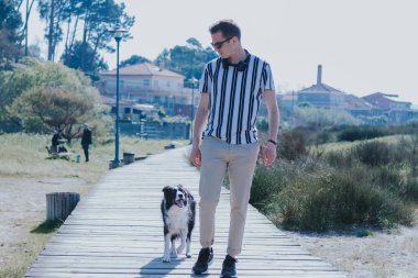 Kulaklıklı bir adam köpeğiyle sahil yolunda yürüyor.