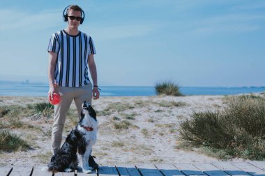 Kulaklıklı ve elinde top olan bir adam sahil yolunda köpeğiyle yürüyor.