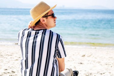 Şapkalı rahat bir adam kumsalda oturmuş denize bakıyor.