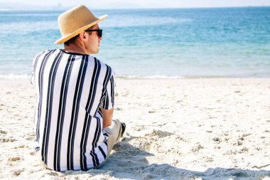 Şapkalı rahat bir adam kumsalda oturmuş denize bakıyor.