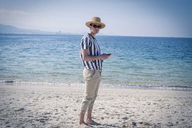 Genç adam plajda cep telefonuyla yürüyor.