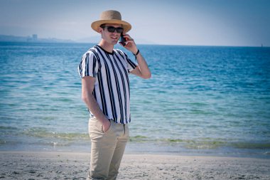 Genç adam plajda cep telefonuyla yürüyor.