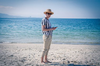 Genç adam plajda cep telefonuyla yürüyor.