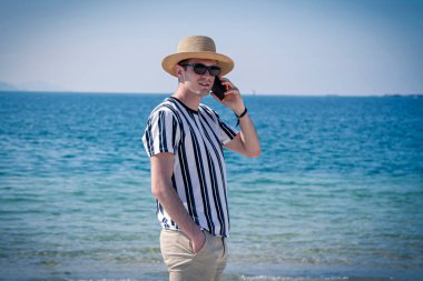 Genç adam plajda cep telefonuyla yürüyor.