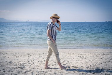 Genç adam plajda cep telefonuyla yürüyor.