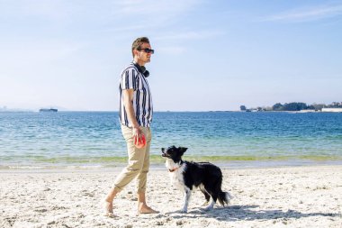 Genç adam sahilde köpek ve topla oynuyor.