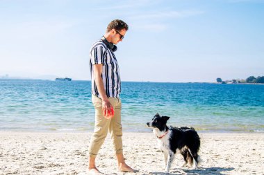 Genç adam sahilde köpek ve topla oynuyor.