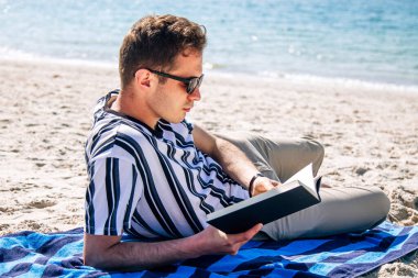 Adam plajda havlusuna uzanmış kitap okuyor.