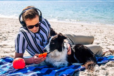 Adam plaj havlusunda uzanmış kulaklıkla müzik dinliyor ve yanında köpeği de var.