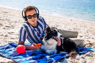Adam plaj havlusunda uzanmış kulaklıkla müzik dinliyor ve yanında köpeği de var.