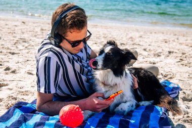 Adam plaj havlusunda uzanmış kulaklıkla müzik dinliyor ve yanında köpeği de var.