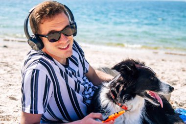 Adam plaj havlusuna uzanmış, kulaklık ve telefonla müzik dinliyor.