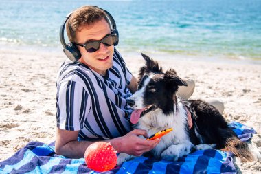 Adam plaj havlusuna uzanmış, kulaklık ve telefonla müzik dinliyor.