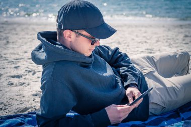 Plajda rahatlamış bir adam dijital tablet ya da e-kitap kullanıyor.