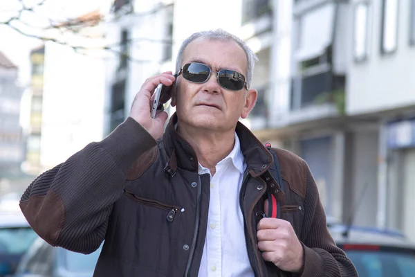 Şık, olgun bir adam sırt çantası ya da çantasıyla şehir kaldırımında yürüyor ve cep telefonunu kullanıyor.