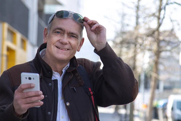 Şık, olgun bir adam sırt çantası ya da çantasıyla şehir kaldırımında yürüyor ve cep telefonunu kullanıyor.