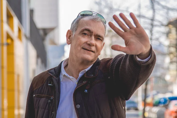 Şık, olgun bir adam kaldırımda yürüyor ve el sallıyor.