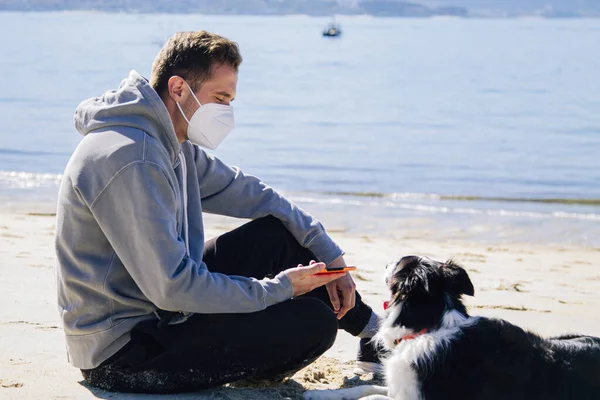 Plajda köpeğiyle maske takan genç bir adam.