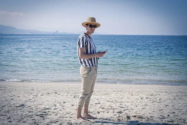 Genç adam plajda cep telefonuyla yürüyor.