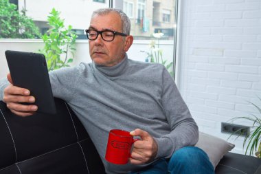 Oturma odasında rahatlamış yetişkin bir adam dijital tablet ya da e-kitap kullanarak bir fincan kahve içiyor.