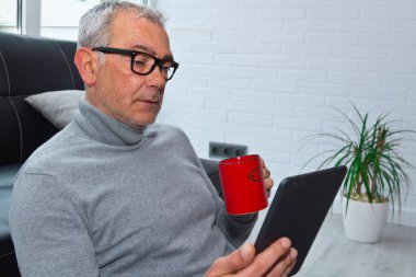 Oturma odasında rahatlamış yetişkin bir adam dijital tablet ya da e-kitap kullanarak bir fincan kahve içiyor.