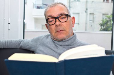 Olgun bir adam evdeki kanepede kitap okuyor.