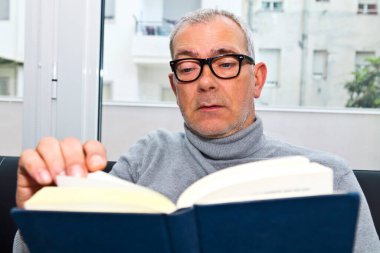 Olgun bir adam evdeki kanepede kitap okuyor.