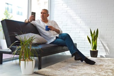 Satisfied man relaxing in his living room using mobile phone while having coffee