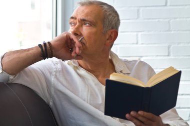 relaxed man reading a book on the armchair and looking out the window