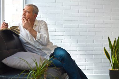 thoughtful mature man writing in notebook on the sofa at home or office