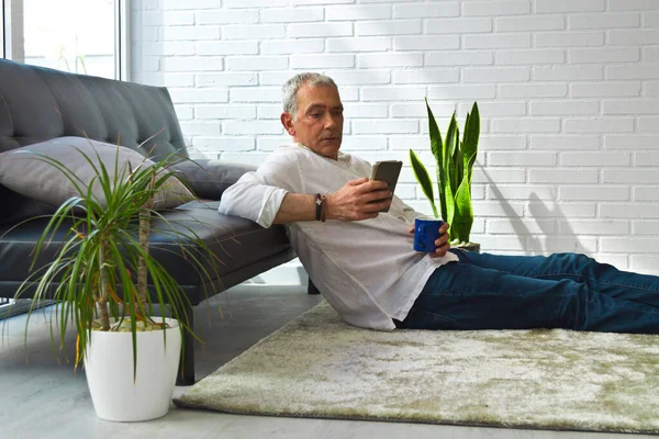 Satisfied man relaxing in his living room using mobile phone while having coffee