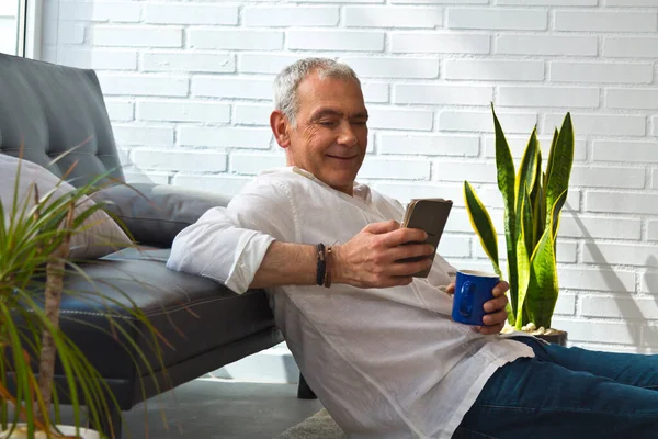 Satisfied man relaxing in his living room using mobile phone while having coffee