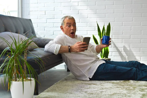 Satisfied man relaxing in his living room using mobile phone while having coffee