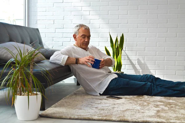 Satisfied man relaxing in his living room using mobile phone while having coffee