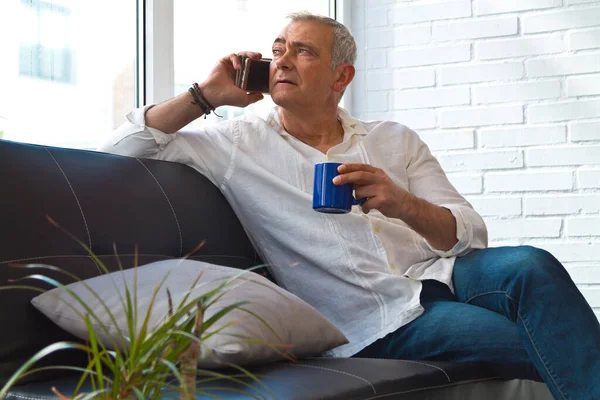 Satisfied man relaxing in his living room using mobile phone while having coffee