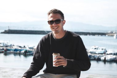 Genç adam iskelede oturuyor ve kahve içiyor.