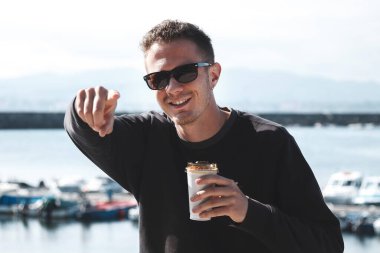 Genç adam gezinti güvertesinde oturuyor, kahve içiyor ve parmağını işaret ediyor.