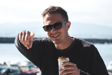 Genç adam iskelede oturuyor, kahve içiyor ve parmağıyla işaret ediyor.