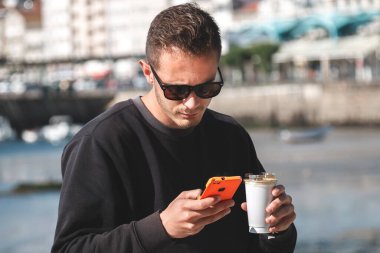 Marinada cep telefonunu kullanırken kahve içen genç bir adam.