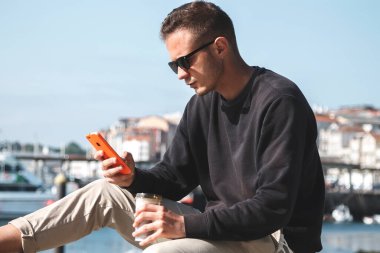 Güneş gözlüklü genç adam hazır kahve içiyor ve arka planda yat limanı olan bir sokakta cep telefonu kullanıyor. Seyahat kavramı