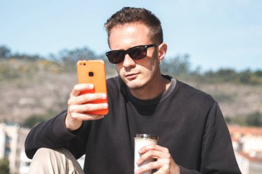 Güneş gözlüklü genç adam hazır kahve içiyor ve arka planda yat limanı olan bir sokakta cep telefonu kullanıyor. Seyahat kavramı