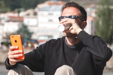 Güneş gözlüklü genç adam hazır kahve içiyor ve arka planda yat limanı olan bir sokakta cep telefonu kullanıyor. Seyahat kavramı