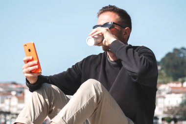 Güneş gözlüklü genç adam hazır kahve içiyor ve arka planda yat limanı olan bir sokakta cep telefonu kullanıyor. Seyahat kavramı