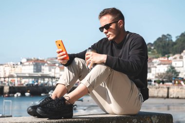 Güneş gözlüklü genç adam hazır kahve içiyor ve arka planda yat limanı olan bir sokakta cep telefonu kullanıyor. Seyahat kavramı