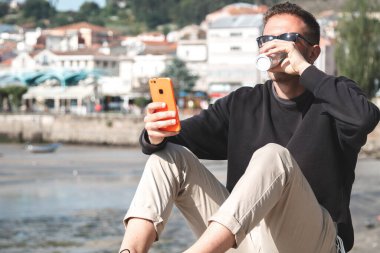 Marinada cep telefonunu kullanırken kahve içen genç bir adam.