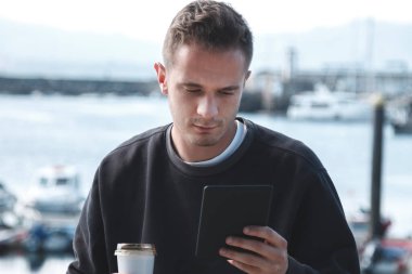 Deniz limanında dijital tablet kullanan ve kahve içip seyahat eden bir adam.