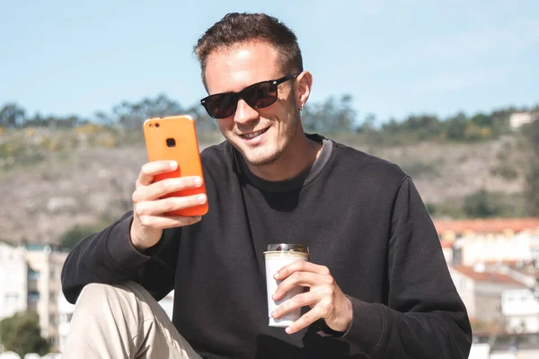 サングラスをかけた若者がテイクアウトコーヒーを飲みながらヨットハーバーを背に路上に座ってる携帯電話を使ってる 旅行のコンセプト — ストック写真