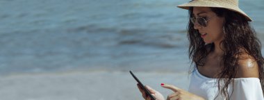 Sahilde şapkalı genç bir kadın dijital tablet ya da e-kitap kullanıyor.