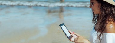 Sahilde şapkalı genç bir kadın dijital tablet ya da e-kitap kullanıyor.